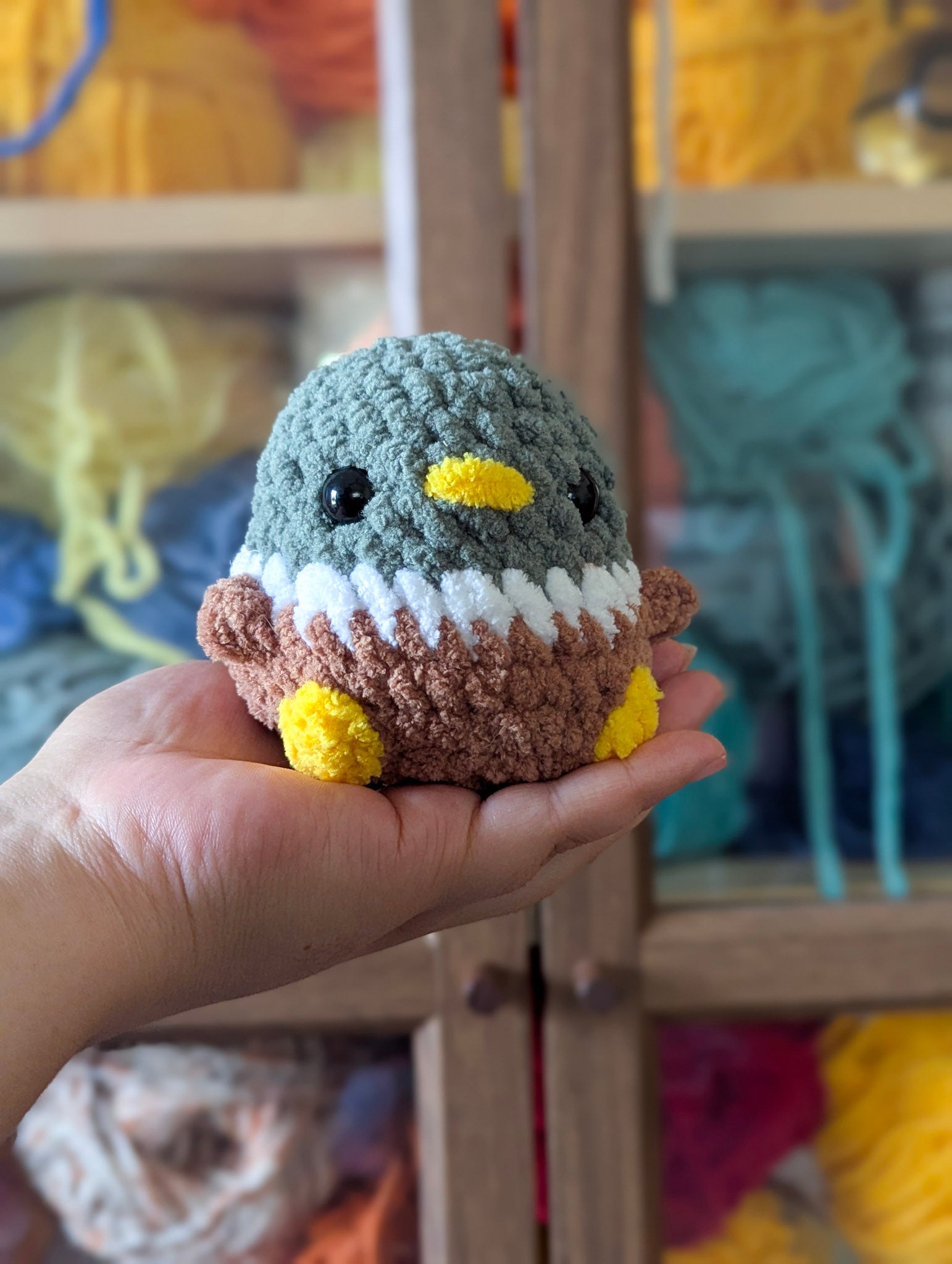 A hand holding a round classic mallard duck in front of a yarn wall.
